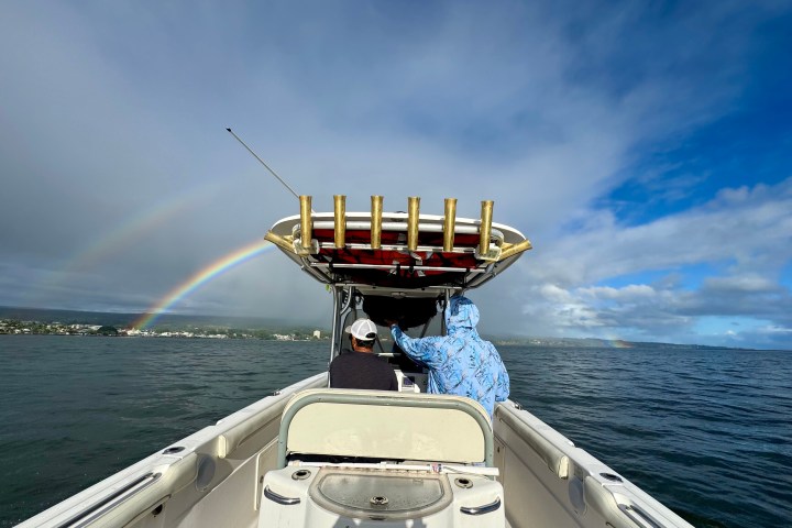 Two people on a boat with a double rainbow over the water and partly cloudy sky.