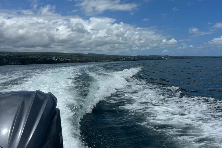 Boat motor and wake on ocean with cloudy sky and distant shoreline.