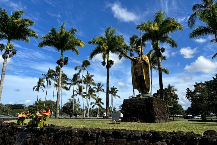 Statue of a figure surrounded by palm trees against a blue sky with clouds.
