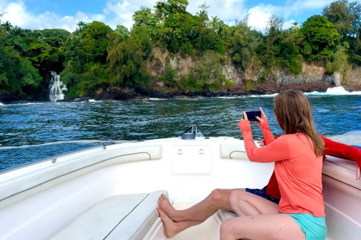 Woman on boat takes photo of waterfall and lush greenery.
