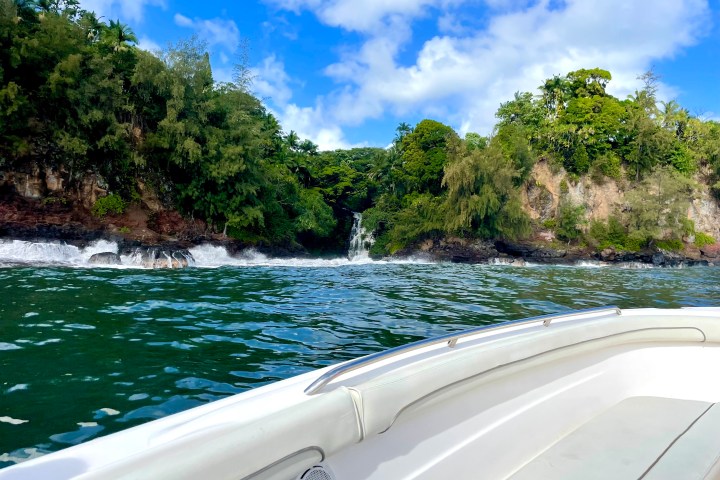 Boat on water approaching lush green cliffs and waterfall under blue sky.