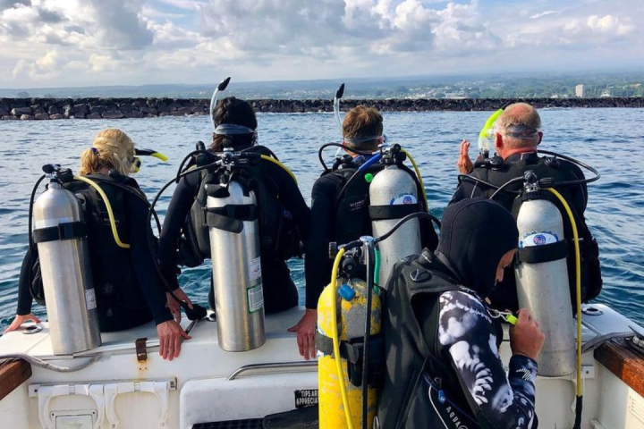 Four scuba divers with tanks sitting on a boat, facing the ocean.