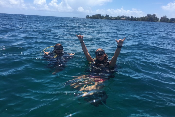 Two scuba divers with masks in ocean, one making shaka signs.