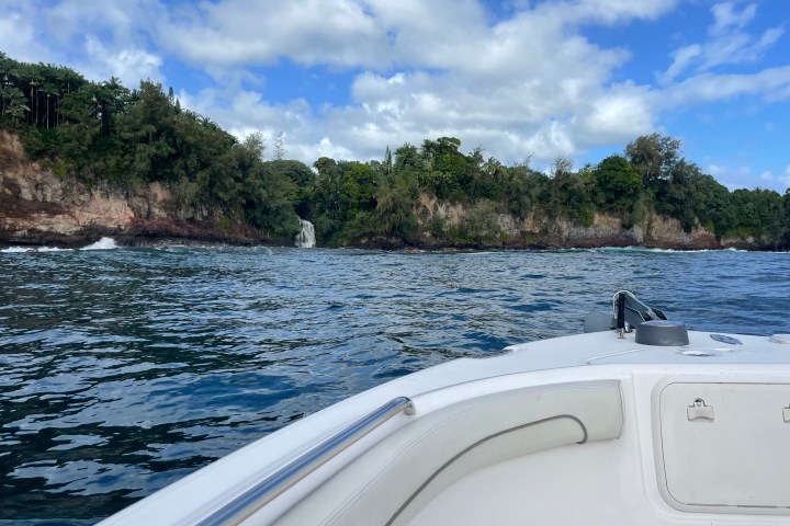 Boat on ocean with lush cliff and waterfall under blue, partly cloudy sky.