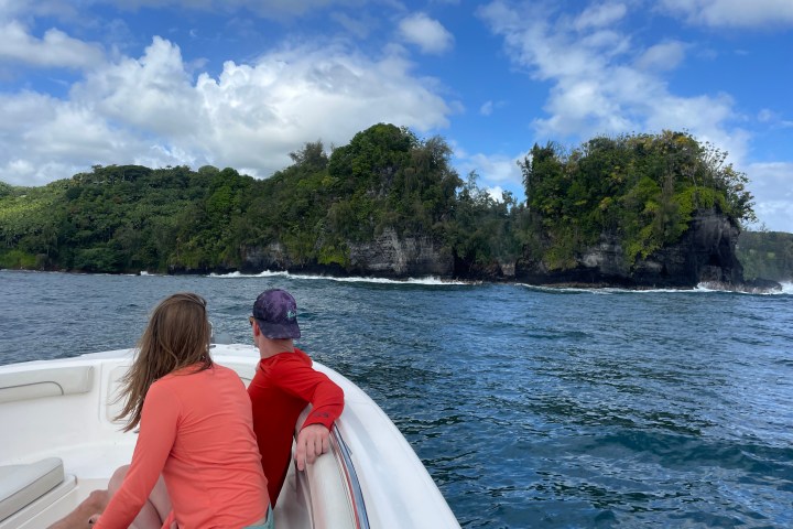 Two people on a boat looking at a lush, rocky coastline with blue sky and clouds.