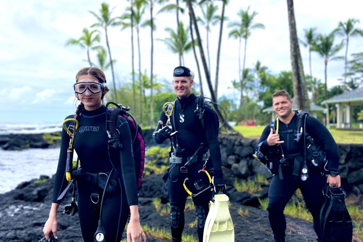 Three divers in wetsuits walk along a rocky shore with palm trees in the background.