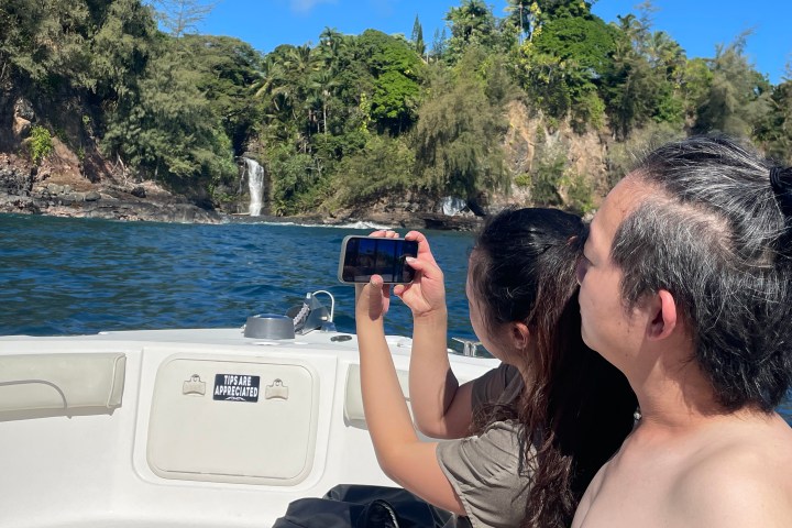 Two people on a boat take a photo of a waterfall and forested cliffs in the distance.