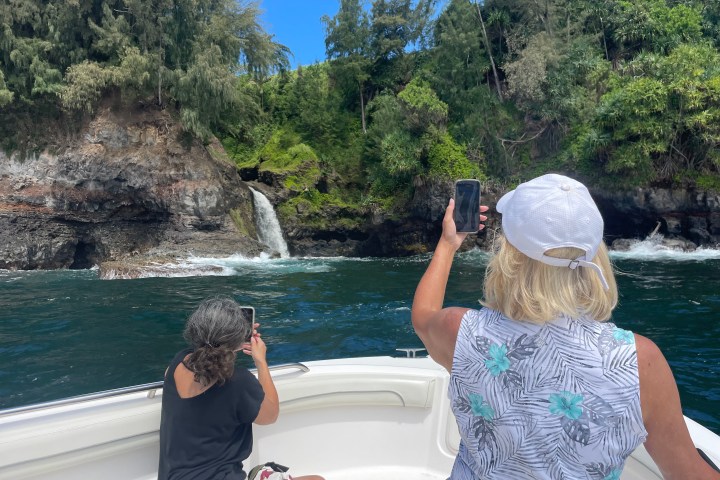 Two people on a boat taking photos of a waterfall on a rocky, forested coastline.