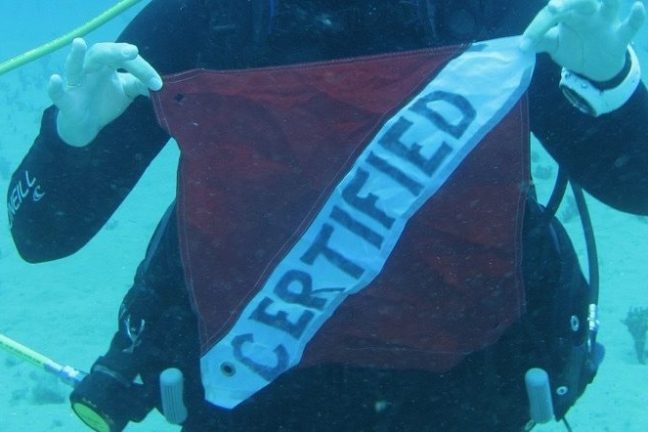 Diver underwater holding a red and white 'CERTIFIED' flag.