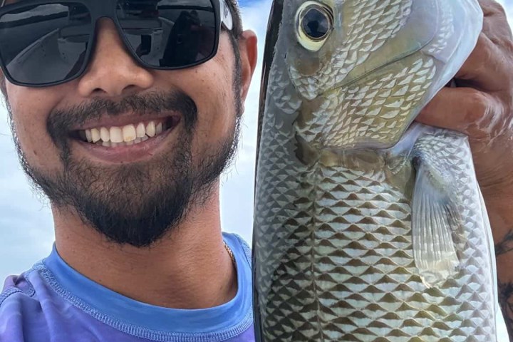 Smiling man with sunglasses and hat holding a fish against cloudy sky.