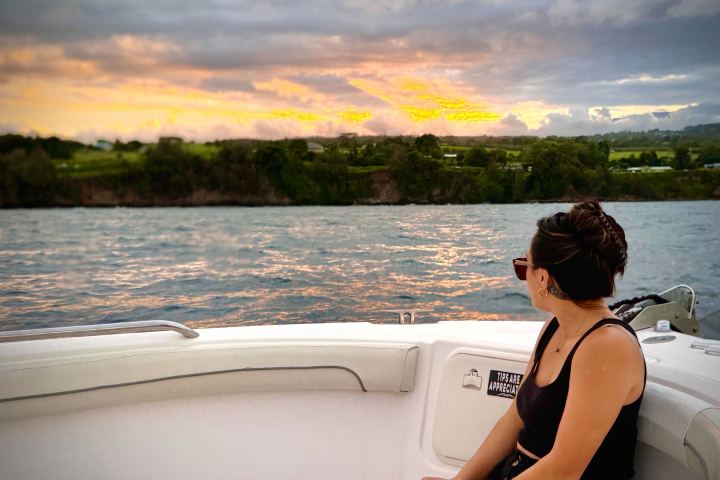 Person relaxing on a boat, watching a colorful sunset over the water and distant trees.