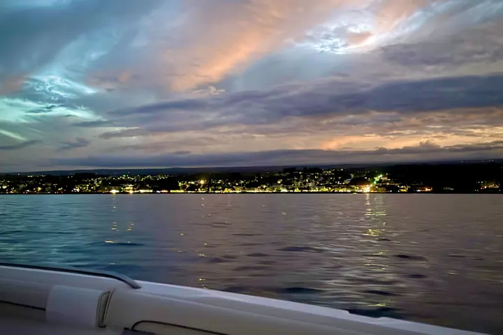 Boat view of illuminated coastal town at dusk with dramatic cloudy sky.