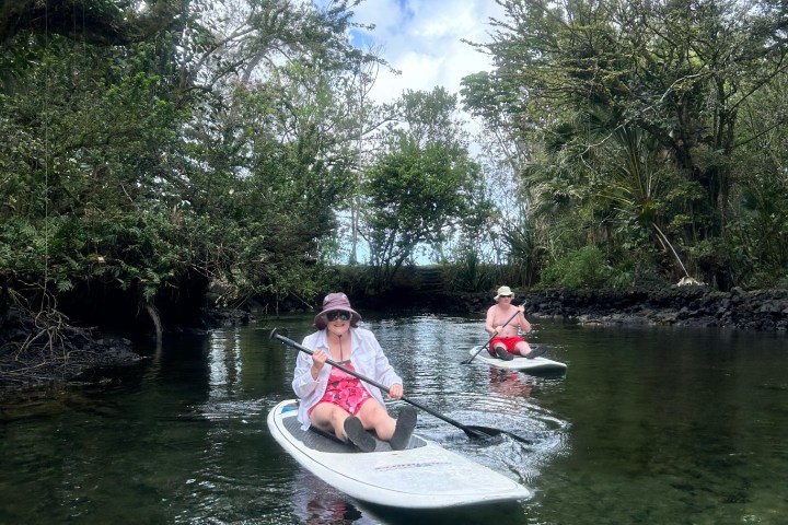 Two people paddleboarding on a narrow river surrounded by lush trees under a partly cloudy sky.