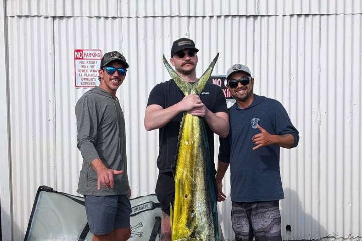 Three men pose with a large fish outside a metal-sided building.