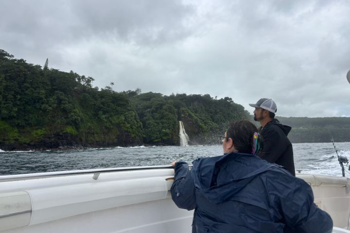 Two people on a boat, looking at a waterfall on a lush green cliff under a cloudy sky.